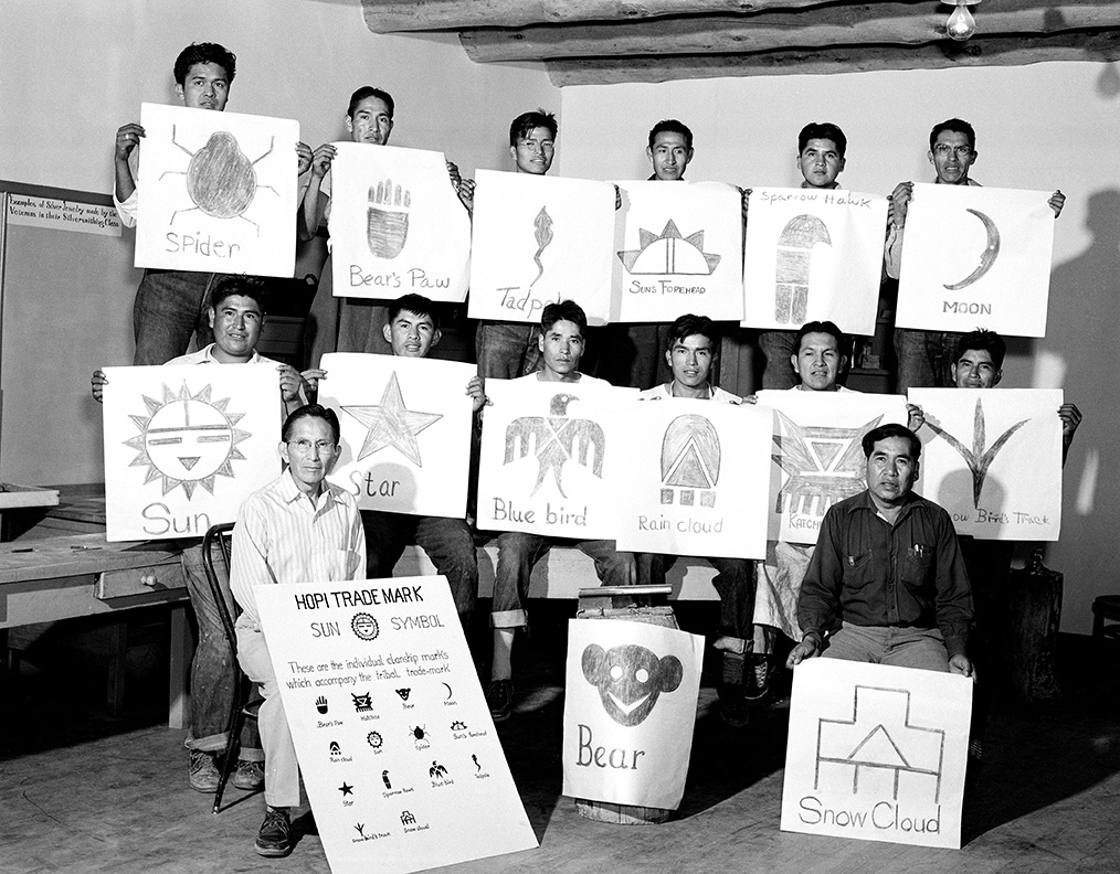 Hopi Silvercraft Cooperative Guild students display drawings of the clan symbols used in their designs. In front of the students are artists and instructors Fred Kabotie (left) and Paul Saufkie.