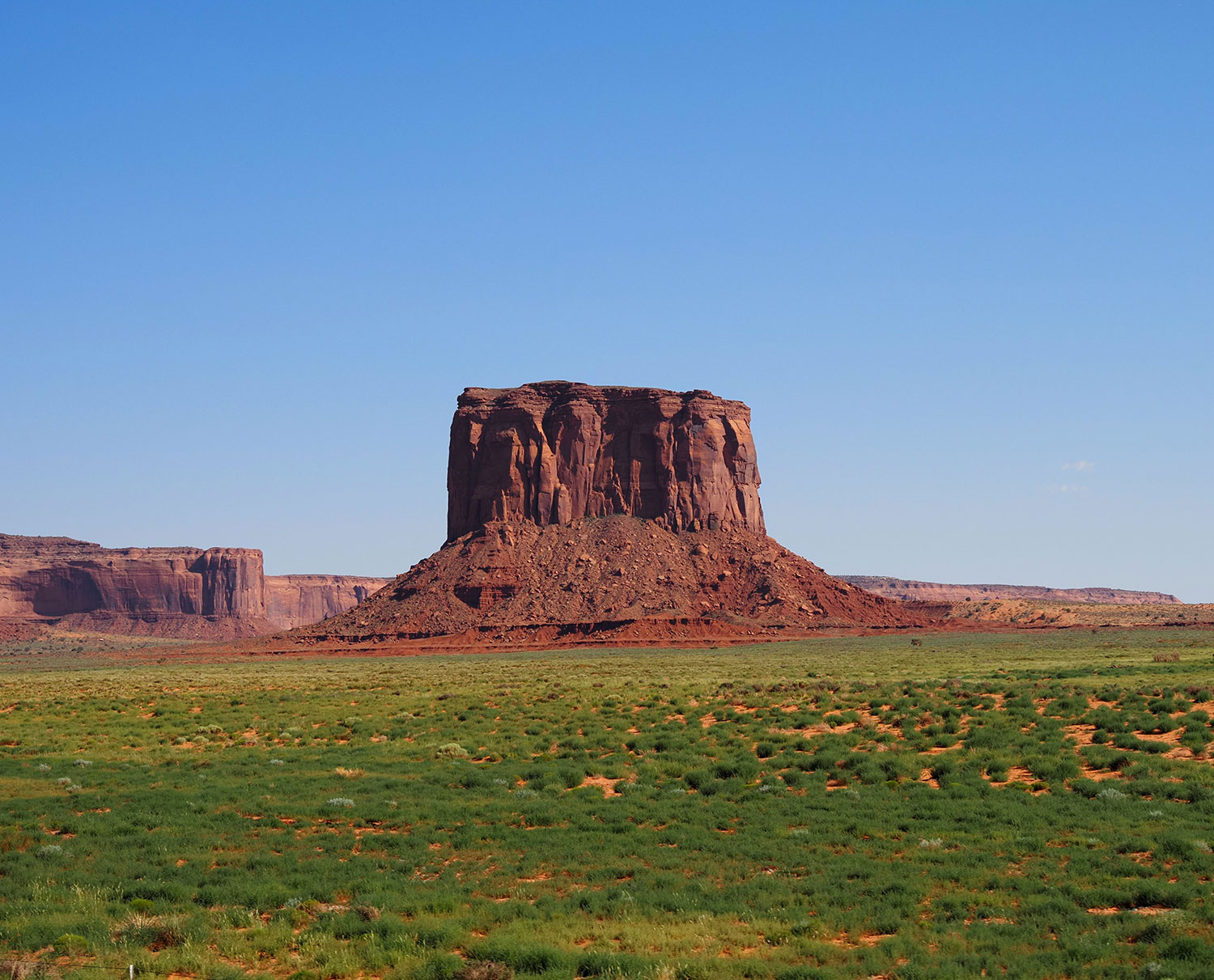 Vibrant vegetation surrounds one of the sandstone formations of Monument Valley. By Michelle Calcagno
