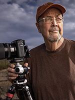 Jack Dykinga poses under a cloudy sky in the desert with a long-lensed camera mounted on a tripod. | JOEL GRIMES 