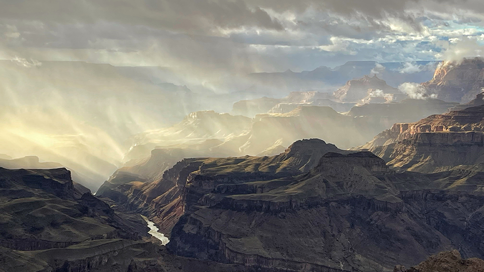 Rays of sunlight slice through clouds at the Grand Cayon, illuminating the background formations and the Colorado River. By Kenton