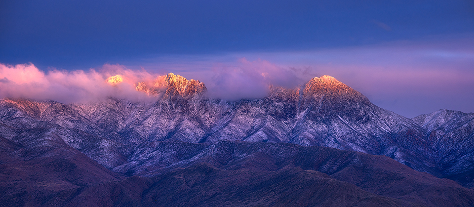 Snow-dusted Four Peaks is partially shrouded in colorful clouds and illuminated by the last rays of light before sunset. By Shakil Chowdhury