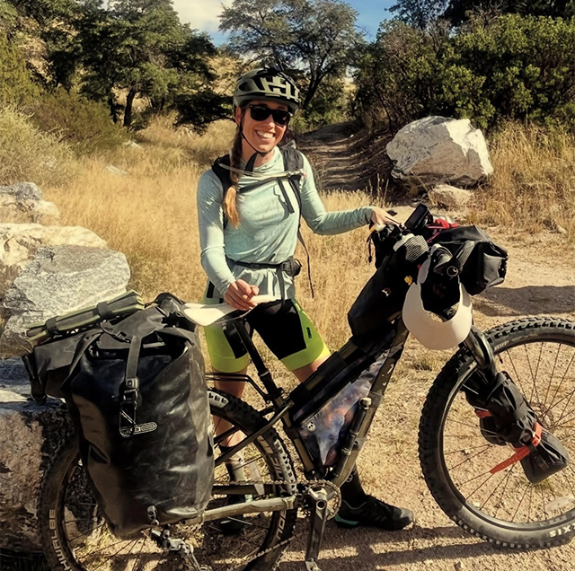Joan Meiners stands next to her bike near some large boulders, grassy hills and small trees along the Arizona Trail. | The Arizona Republic