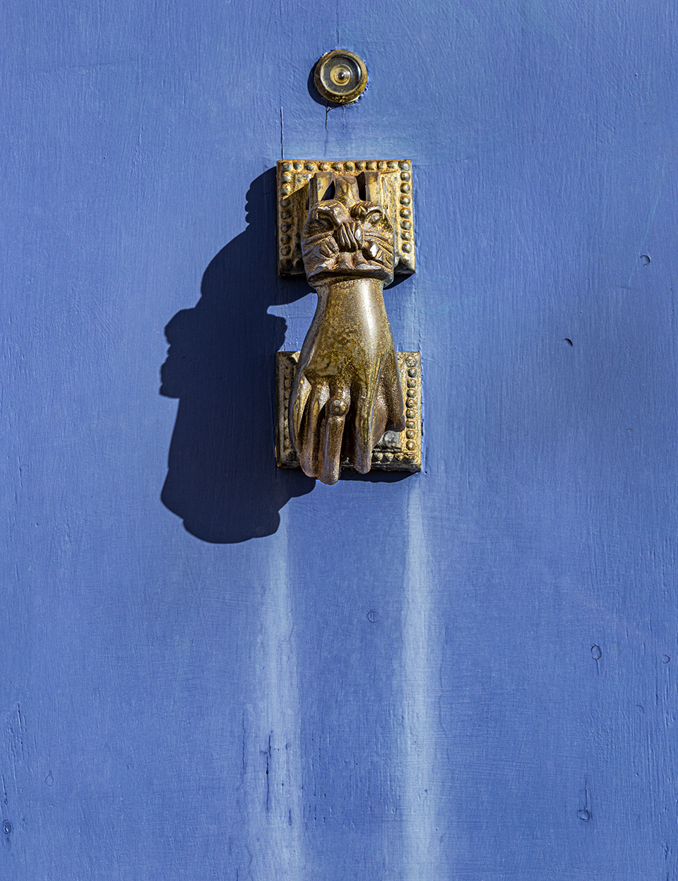 A metal door knocker in the shape of a human hand stands out against its blue backdrop. By Steven Meckler