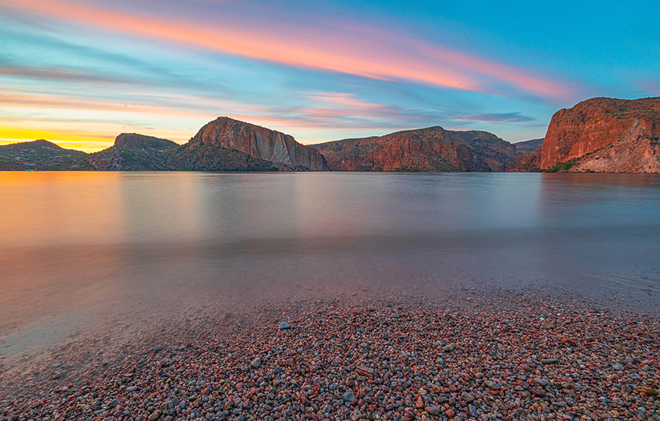A serene sunset at Canyon Lake creates a colorful texture of pebbles at the water's edge. By Hasanur Khan