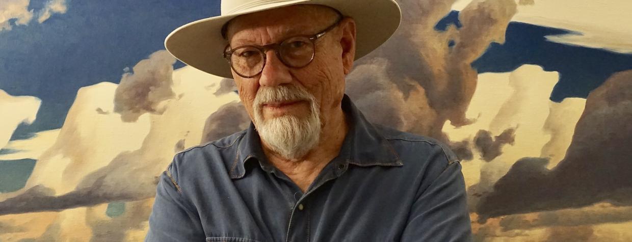 Painter Ed Mell, in a blue shirt and cream colored-hat, stands with his arms crossed in front of one of his landscape paintings. 