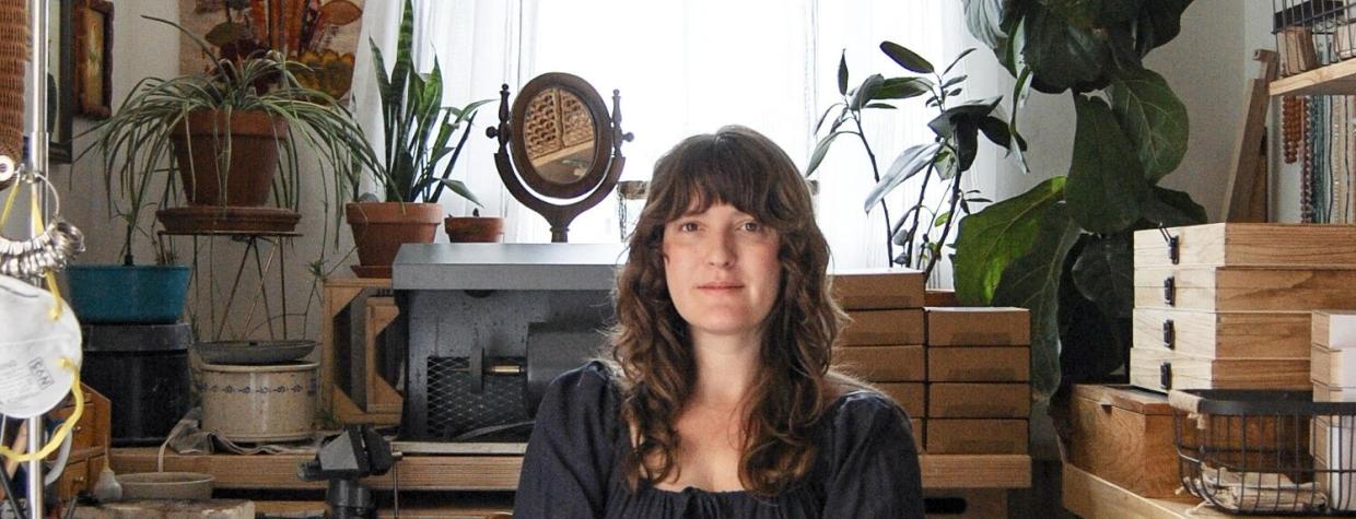 A woman with long hair sits in a dark dress in her jewelry studio. Boxes of gems, houseplants and other ephemera  surround her. 