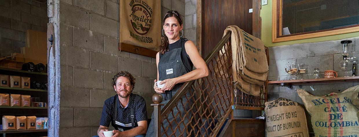 Daniel and Monica Garland pose with cups of coffee on a staircase near sacks of coffee beans. By David Zickl
