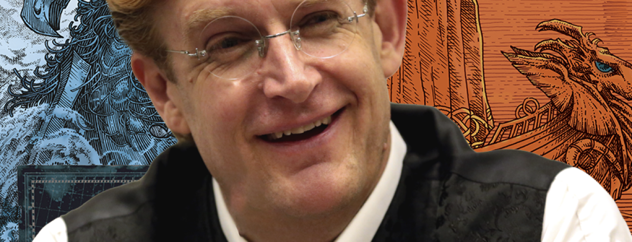 A middle-aged white man in glasses and a black vest poses in front of an illustration of one of his comic book characters