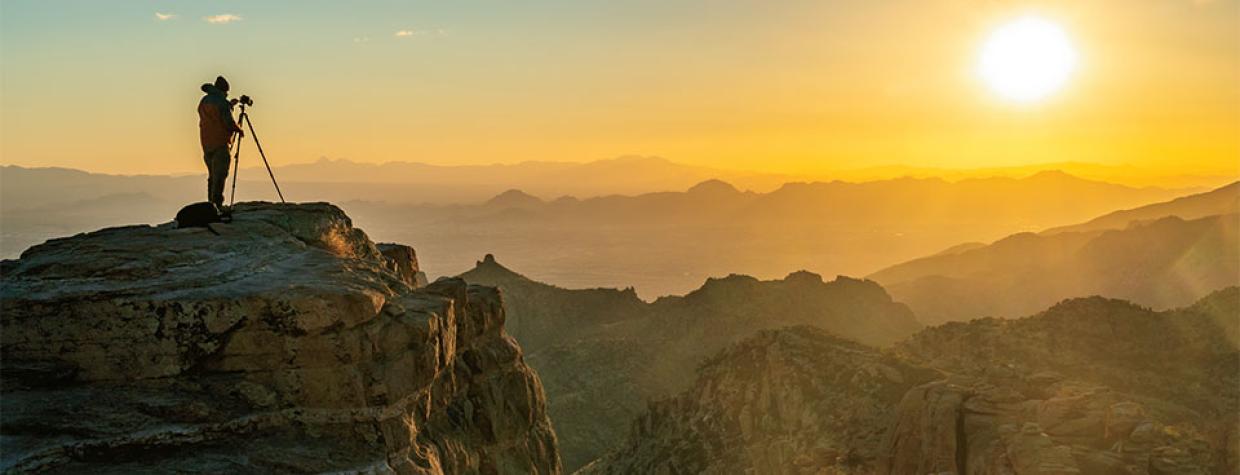 Photographer with tripod standing on rock cliff looking toward sunset.