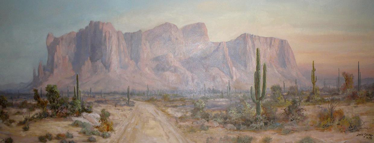 Superstition Mountain is the title of this painting by David Swing, depicting the iconic mountain with a foreground of a dirt road through desert landscape dotted by saguaros.
