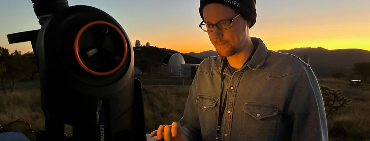 A young man in a black beanie and glasses calibrates a large telescope.