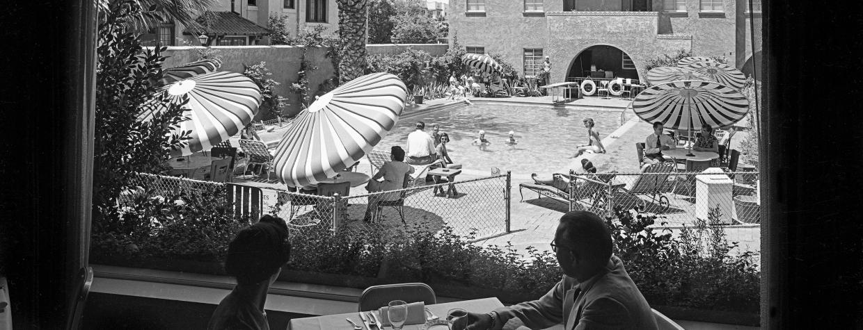 A black and white image shows two people, a woman and a man, in shadow at a dining table within the interior of the Westward Ho hotel in downtown Phoenix. The pair looks out onto a busy swimming pool, surrounded by striped sunbrellas. 