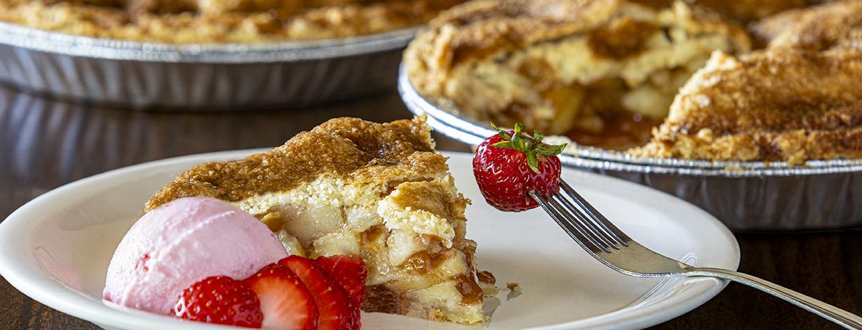 A slice of pie with strawberry slices and ice cream is plated along side several whole pies. By Steven Meckler