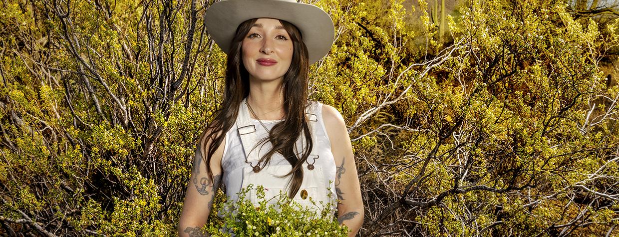 Stina Swesey poses in a desert setting holding a bunch of creosote flowers. By Steven Meckler