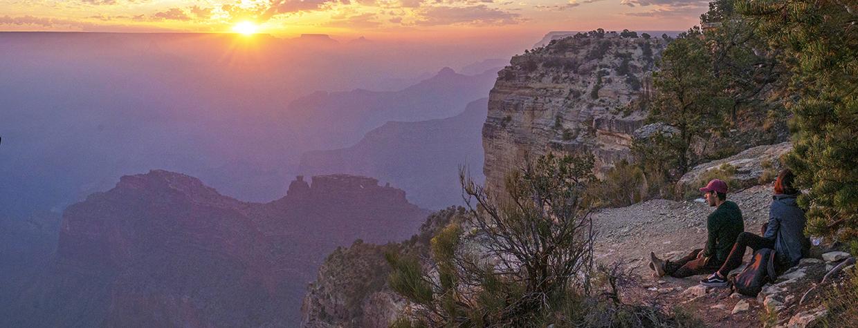 Two people watch the sun set from the edge of the Grand Canyon.