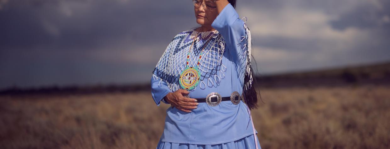 An indigenous woman wears periwinkle blue traditional clothing and runs her hand through her hair. She is against a monsoonal backdrop and standing in a field of dead grass. 