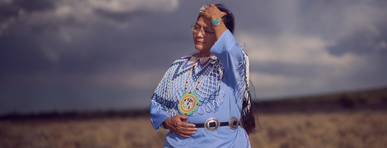 An indigenous woman wears periwinkle blue traditional clothing and runs her hand through her hair. She is against a monsoonal backdrop and standing in a field of dead grass. 