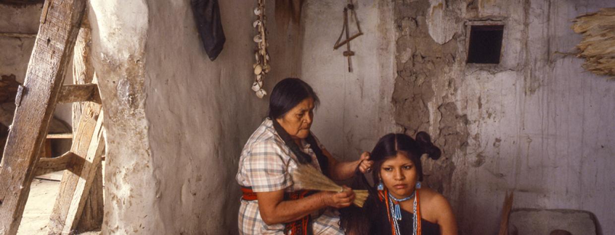 One indigenous woman sits on a crate, fixing another indigenous woman's hair. 