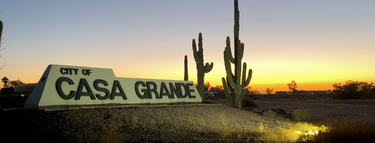 A sign welcoming visitors to Casa Grande is framed by saguaros and lit by spotlights.