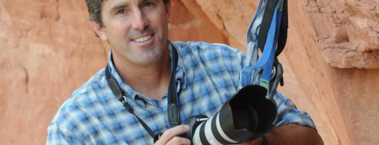 A color photograph shows a smiling white man in a blue plaid shirt. He is anchored to a rock wall with climbing gear and holding a camera with a large lens. 