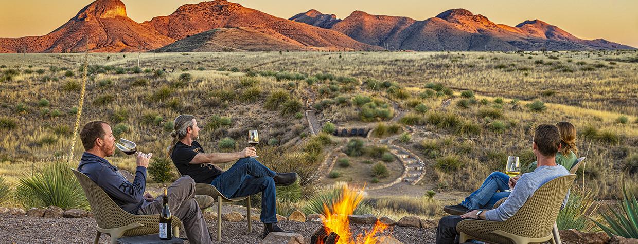 Photograph of four people sitting around a fire at dusk with a view of the nearby Santa Rita Mountains is by Steven Meckler.