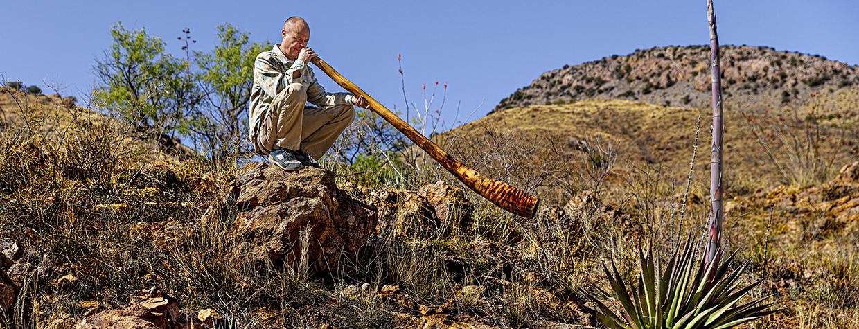 Photograph of man crouching on a rock in the desert blowing into a didgeridoo is by Steven Meckler