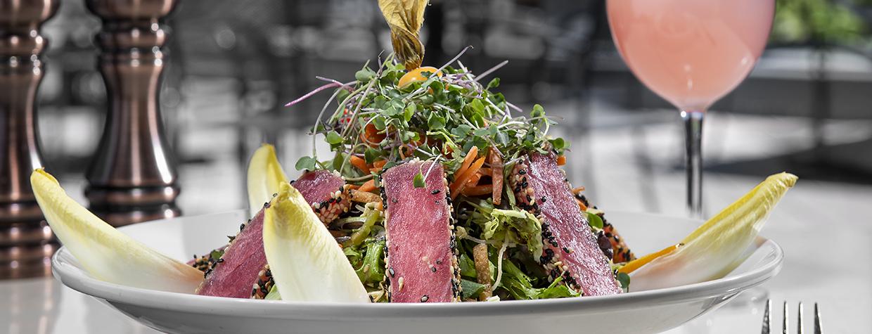 Photograph of an artistic salad and a pink drink garnished with flowers against a soft backdrop of a red rock view is by Rob Ballard.
