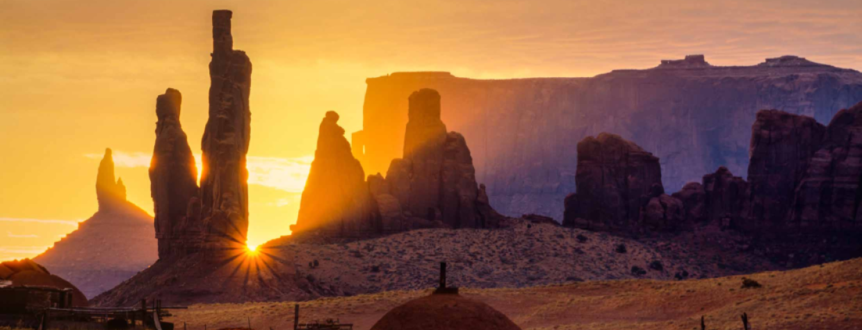 The sun peeks between the iconic spires of Monument Valley Navajo Tribal Park. By David Muench