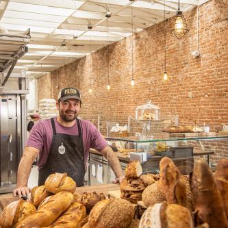 Jonathan Przybyl, dressed in apron at his shop, with loaves of bread and other baked goods.