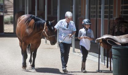 A trainer and young client walk alongside a dark brown horse. 