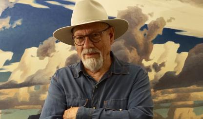 Painter Ed Mell, in a blue shirt and cream colored-hat, stands with his arms crossed in front of one of his landscape paintings. 