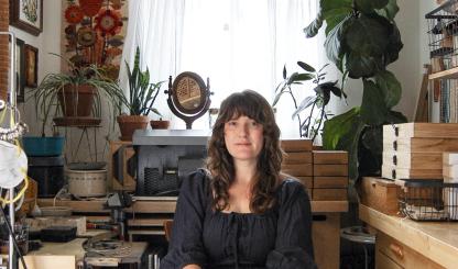 A woman with long hair sits in a dark dress in her jewelry studio. Boxes of gems, houseplants and other ephemera  surround her. 