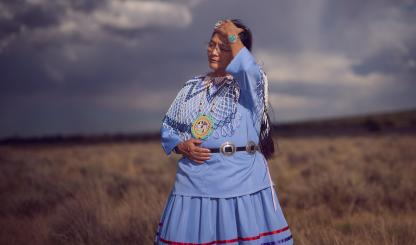 An indigenous woman wears periwinkle blue traditional clothing and runs her hand through her hair. She is against a monsoonal backdrop and standing in a field of dead grass. 