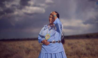 An indigenous woman wears periwinkle blue traditional clothing and runs her hand through her hair. She is against a monsoonal backdrop and standing in a field of dead grass. 