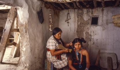 One indigenous woman sits on a crate, fixing another indigenous woman's hair. 