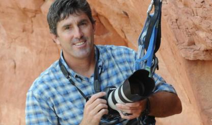A color photograph shows a smiling white man in a blue plaid shirt. He is anchored to a rock wall with climbing gear and holding a camera with a large lens. 