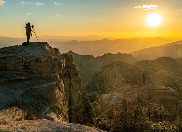 Photographer with tripod standing on rock cliff looking toward sunset.