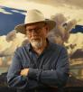 Painter Ed Mell, in a blue shirt and cream colored-hat, stands with his arms crossed in front of one of his landscape paintings. 