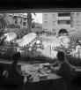 A black and white image shows two people, a woman and a man, in shadow at a dining table within the interior of the Westward Ho hotel in downtown Phoenix. The pair looks out onto a busy swimming pool, surrounded by striped sunbrellas. 
