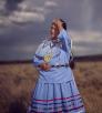 An indigenous woman wears periwinkle blue traditional clothing and runs her hand through her hair. She is against a monsoonal backdrop and standing in a field of dead grass. 
