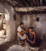 One indigenous woman sits on a crate, fixing another indigenous woman's hair. 