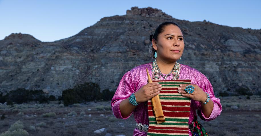 A Diné weaver holds weaving tools before a red-rock mesa