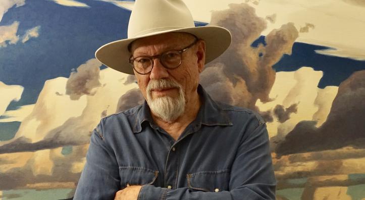 Painter Ed Mell, in a blue shirt and cream colored-hat, stands with his arms crossed in front of one of his landscape paintings. 
