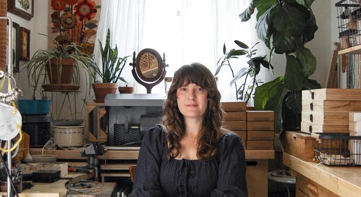 A woman with long hair sits in a dark dress in her jewelry studio. Boxes of gems, houseplants and other ephemera  surround her. 