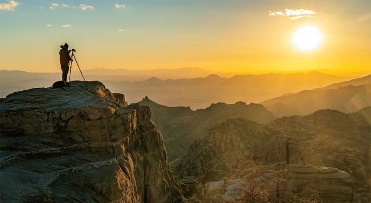 Photographer with tripod standing on rock cliff looking toward sunset.