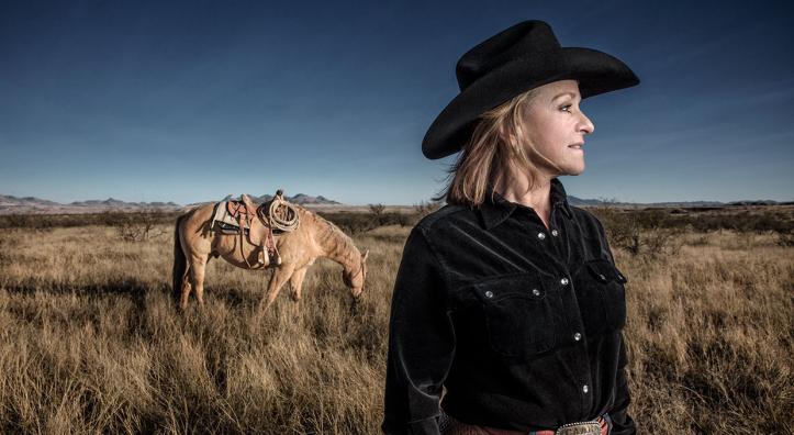 Deborah Copenhaver Fellows looks off into the distance standing in tall grass with her horse a few steps away. By Joel Grimes