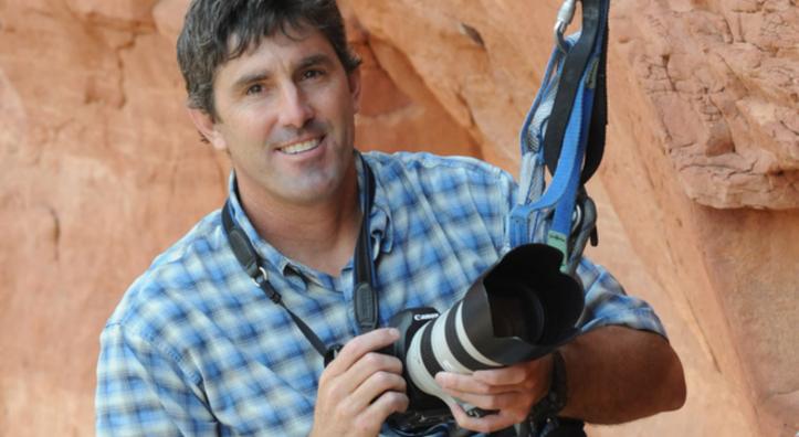 A color photograph shows a smiling white man in a blue plaid shirt. He is anchored to a rock wall with climbing gear and holding a camera with a large lens. 