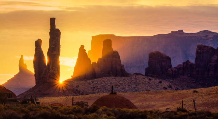 The sun peeks between the iconic spires of Monument Valley Navajo Tribal Park. By David Muench