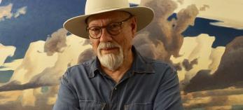 Painter Ed Mell, in a blue shirt and cream colored-hat, stands with his arms crossed in front of one of his landscape paintings. 