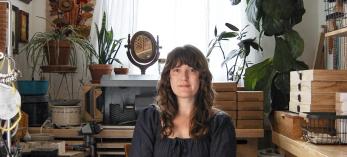 A woman with long hair sits in a dark dress in her jewelry studio. Boxes of gems, houseplants and other ephemera  surround her. 